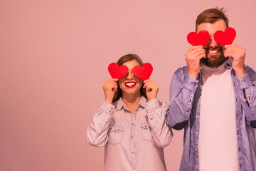 Cheerful multiracial happy couple covering eyes with red paper hearts and smiling pink background