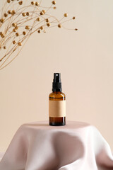 Brown amber glass spray bottle with label mockup and dry flowers on dais podium on beige background.