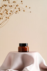 Brown amber glass jar of natural moisturizing face cream on podium with dry flowers on beige...