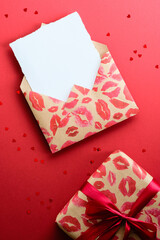 Craft paper envelope with lipsticks marks and gift box on red background with heart shaped confetti. Happy Valentine's Day concept. Flat lay, top view.