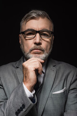 senior pensive businessman in eyeglasses touching chin while looking at camera isolated on black.