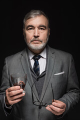 stylish senior man with glass of whiskey and eyeglasses looking at camera isolated on black.