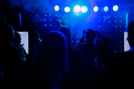 cheering crowd in front of bright blue stage lights. Silhouette image of people dance in disco night club or concert at a music festival.
