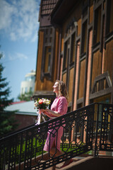 Fototapeta na wymiar Young happy blonde woman in a pink dress with a large bouquet of flowers walks around the old European city and enjoys life on a summer day, beautiful portraits with a huge bouquet of flowers.