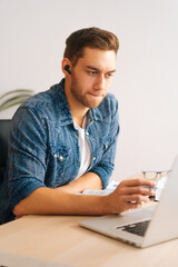 Vertical shot of serious young business man in earphone having online video conference on laptop at home office. Confident male student watch webinar training course on computer sitting at desk.