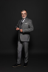 full length view of happy senior man in suit looking at camera on dark grey.