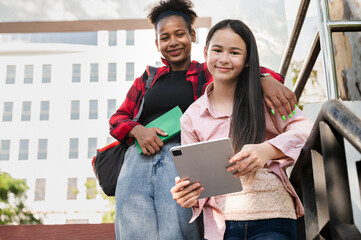 Happy girl student holding tablet and book with friend and looking camera at school park