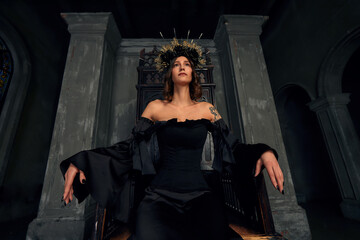 Portrait of a beautiful medieval young woman in black dress In stylish crown sitting on a throne....