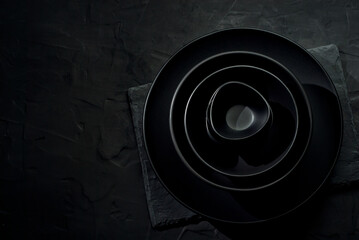 Obraz premium Black tableware on a dark background. Plates and bowls of different shapes and sizes are stacked in a pile. Contrasting dramatic light as an artistic effect.