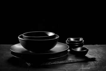 Black tableware on a dark background. Plates and bowls of different shapes and sizes are stacked in a pile. Contrasting dramatic light as an artistic effect.