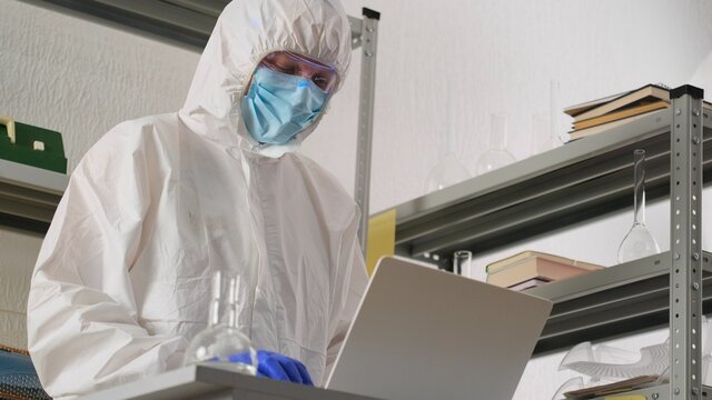 Man scientist or medical working, in protective uniform, looking on flask with liquid in science lab and recording results to laptop