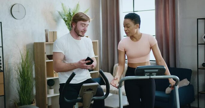 Home workout concept where attractive smiling sportive multiethnic couple exercising together on treadmill and exercise bike and talking about joint photos on guy's mobile