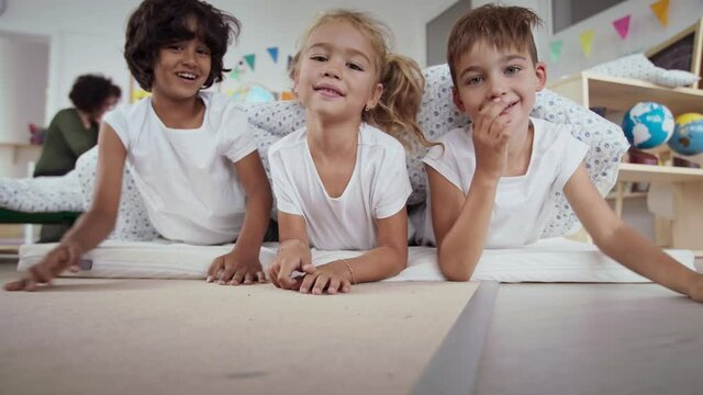 Preschool children in pajamas having fun hiding under blanket indoors in nursery school.