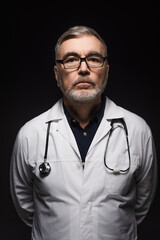 senior bearded doctor with stethoscope looking at camera isolated on black.