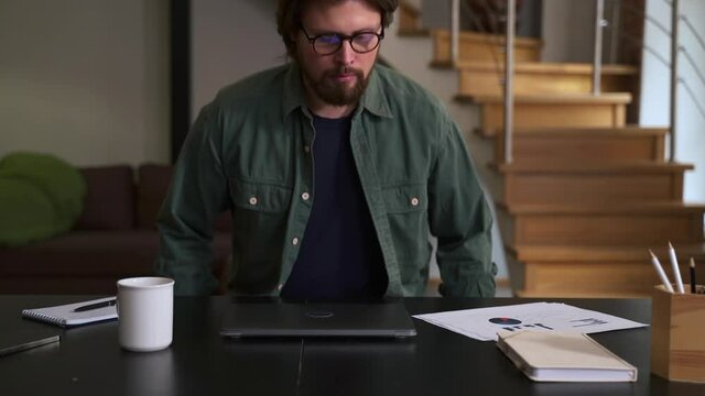 Young man sits down at desk and opens laptop to working in home office spbas. Closeup view of bearded American businessman starts work and looks at computer screen, sits at table in light interior