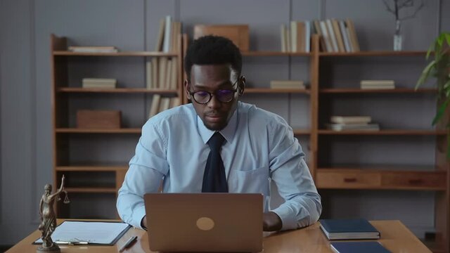 African American businessman working with computer and posing, sitting at table in office spbas. Front view of young man opens laptop and looks at screen, poses with smile and sits at desk with statue