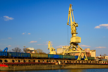 River port crane loading open-top gondola cars on sunny day. Empty river drag boats or barges moored by pier, Empty cars ready for loading.