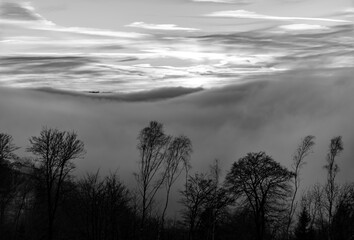 Sauerland sunset panorama on a foggy winters evening. Cloudy evening sky, wafts of mist in Lenne valley near Altena Germany. Mystic atmosphere in rural landscape, black and white greyscale.