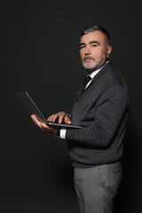 senior bearded man in jumper holding laptop and looking at camera on dark grey.