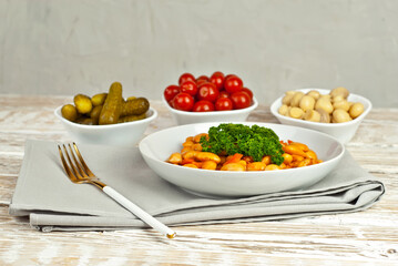 Beans with carrots in a white bowl. Beans with pickled tomatoes, mushrooms and cucumbers on a white background. Food on a shabby table. Bowl on an old wooden board.