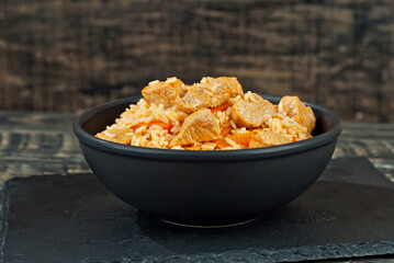 Pilaf with meat and carrots in a black bowl. Pilaf on a black background. Food on a shabby table. Bowl on an old wooden board.