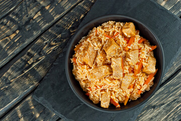 Pilaf with meat and carrots in a black bowl. Pilaf on a black background. Food on a shabby table. Bowl on an old wooden board. Copy space and free space for text near food.