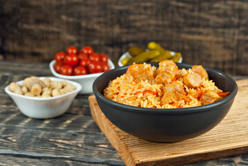 Pilaf with meat and carrots in a black bowl. Marinated mushrooms, tomato and cucumber on a black background. Food on a shabby table. Bowl on an old wooden board.