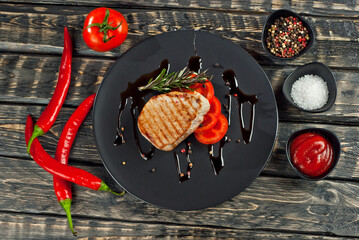 Steak with sliced tomatoes in a black plate. Meat on a dark wooden background. Food, sauce, salt, pepper and spices on an old shabby table.