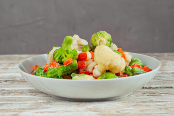 A mixture of frozen vegetables in a white bowl. Preparation for a dish on an old shabby table. Sliced vegetables on a light board background.