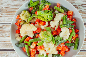 A mixture of frozen vegetables in a white bowl. Preparation for a dish on an old shabby table. Sliced vegetables on a light board background.