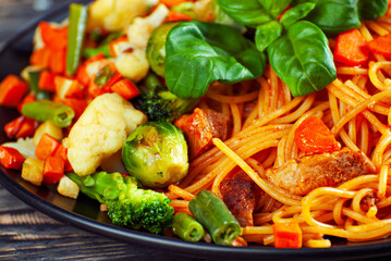 Noodles with carrots, vegetables and meat in a black plate. Food on a black Yerevan table. Basil leaves on pasta. Pasta with a mixture of vegetables.