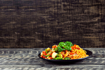 Noodles with carrots, vegetables and meat in a black plate. Food on a black Yerevan table. Basil leaves on pasta. Pasta with a mixture of vegetables. Copy space and free space for text next to food.