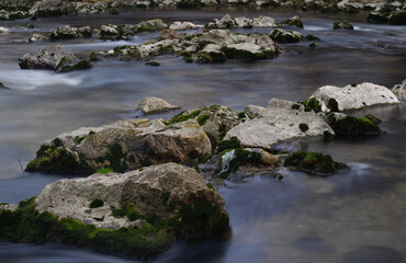 Large stones in the river lie side by side and one behind the other. The water flows around the stones in a long exposure