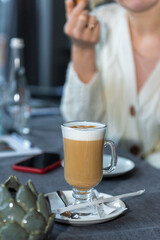 a glass of latte on a saucer. A girl is sitting at the table. face is not visible