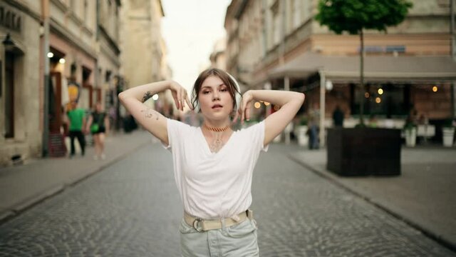 Outdoor summer lifestyle portrait of young pretty hipster woman in wireless headphones having fun, listening music and dancing on the street. Beautiful sexy woman with tattoo dancing on a street