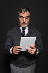 surprised senior man in jumper and tie looking at digital tablet isolated on dark grey.