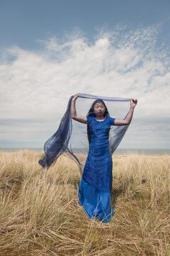 Young woman in blue dress in dunes near ocean holding thin fabric