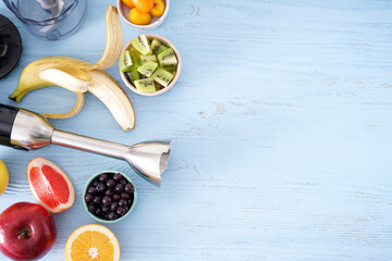 Electric hand blender with stainless steel body with accessories and fruits on wooden background, place for text. Top view.