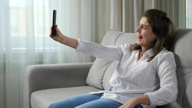 Cheerful disabled woman with cerebral palsy takes selfie via mobile phone smiling and making gestures sitting on comfortable sofa at home.