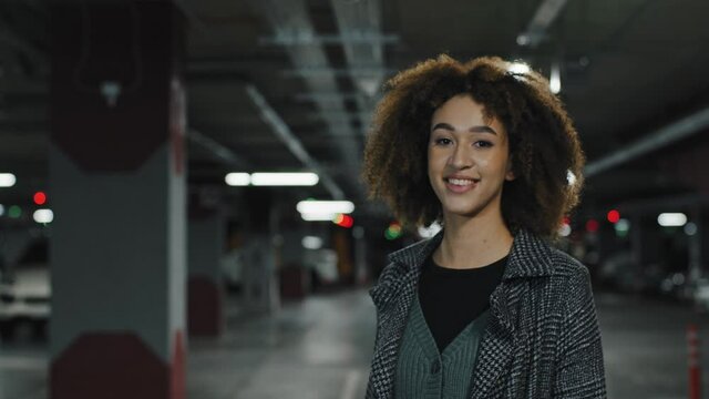 View From Behind Beautiful Young Girl Business Woman Lady African American Female With Curly Hair Stylish Hairstyle In Gray Coat Walks In Parking Lot Turns Around Looking At Camera Smiling Friendly