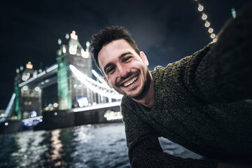 Happy young man taking selfie in Tower Bridge, London - Handsome guy smiling at camera on city street - Tourism and happy lifestyle concept