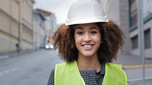 Female portrait worker profession close-up african american woman girl with curly hair civil engineer professional wearing safety helmet standing in city street and looking at camera smiles friendly