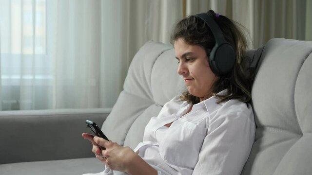 Disabled woman with infantile cerebral paralysis in headphones listens to music singing alone and holding phone sitting on sofa at home closeup.