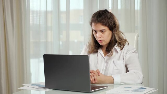 Disabled woman freelancer with infantile cerebral paralysis talks on videocall via laptop with manager showing project paper against window at home.