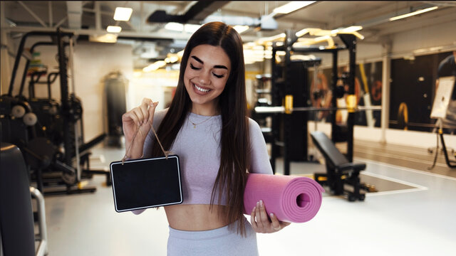 Beautiful young woman holding yoga mats and a empty sign in her gym. Reopening after covid-19 lockdown concept. Fitness club owner holding wooden sign in her hands, standing in front of the gym.