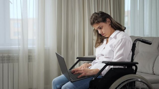 Young disabled long haired woman with cerebral palsy works online remotely via laptop sitting in wheelchair near window in living room at home.