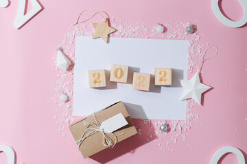 2022 number on cubes over a blank sheet of paper surrounded by new year decoration. Artificial plastic snow, toy stars, present box. All arranged in a composition. Over pink