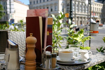 Summer outdoor terrace in a cafe in a large European city, on a warm sunny day. A white cup of coffee, water, menu, sugar bowl, pepper shaker on the table. Lifestyle scene. Tourist's breakfast.