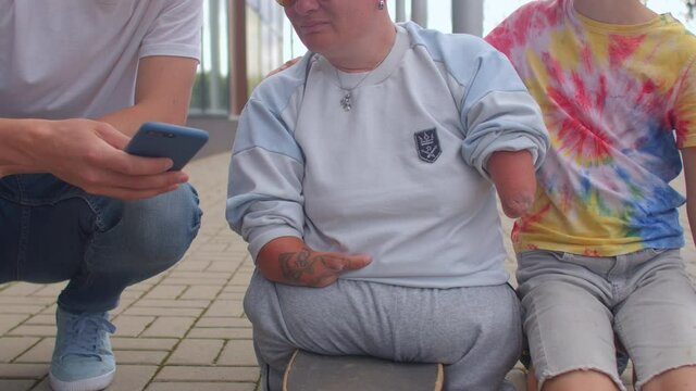 Woman with congenital malformations of the upper and lower endpoints on a skateboard surrounded by friends using the phone.