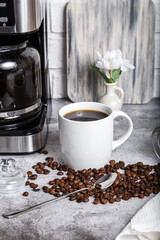 A black drip electric coffee machine with a glass teapot brews a morning drink. Household appliances, a white cup and a jar of beans on the kitchen table on the countertop at home